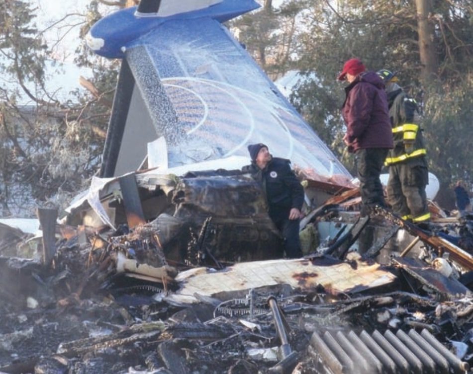 The burnt wreckage of the Colgan Air Q400, its tail section lying on the debris, with people in winter clothing and a firefighter on the site.