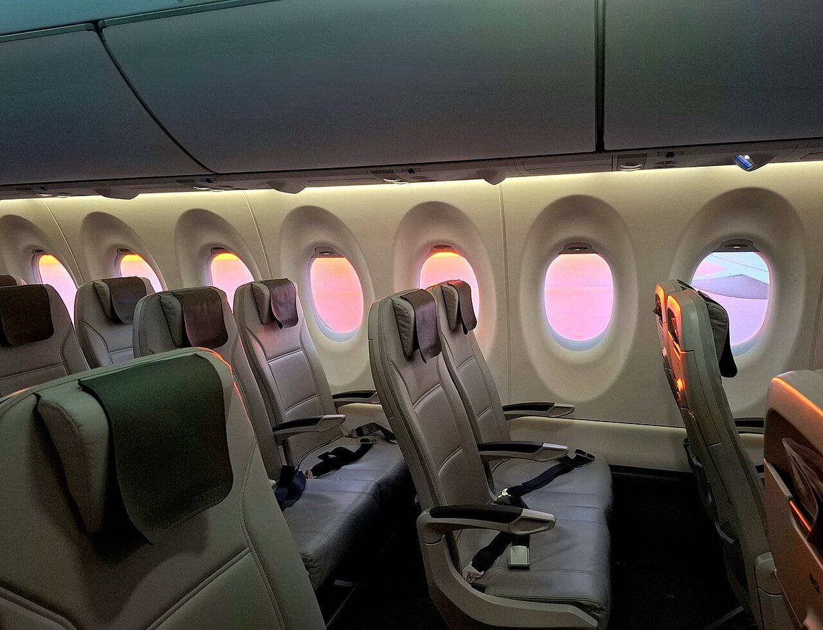 An airliner cabin seen from the aisle: rows of grey leather seats, large oval windows glowing pink at sunrise and closed overhead bins.
