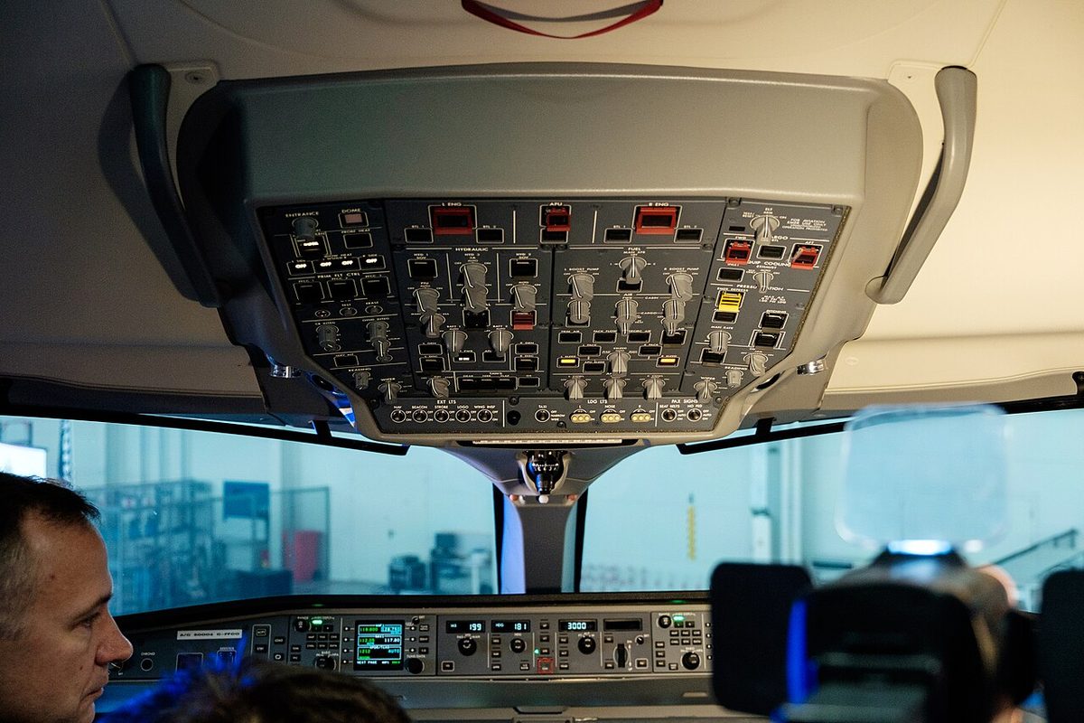 The overhead panel of an airliner flight deck: three switches under red guards across the top, hydraulic and fuel panels in the middle and a row of light switches along the bottom.
