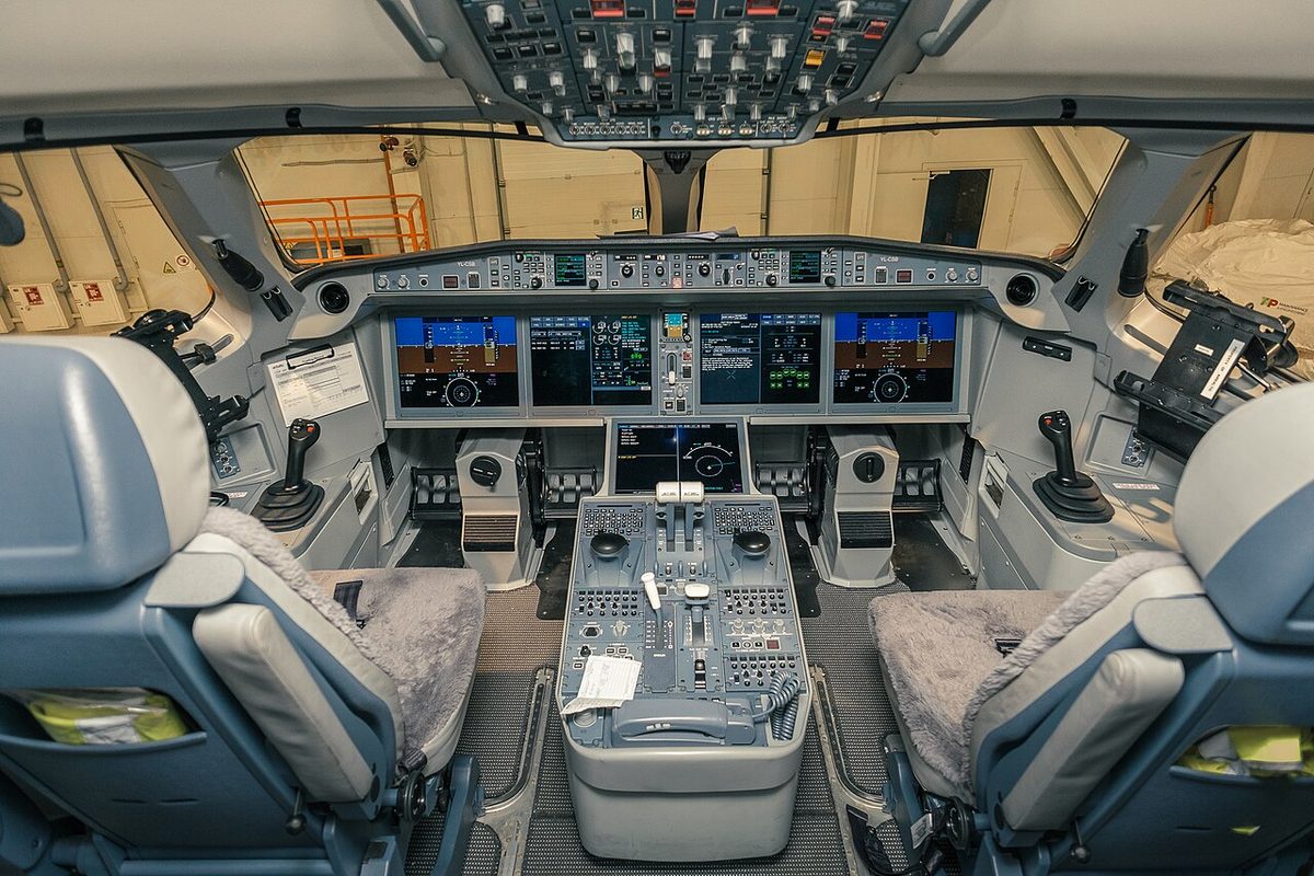 An airliner flight deck in a hangar seen from between the seats: a small side-stick on each pilot's outboard side console, four large display screens across the main panel, a fifth at the front of the centre pedestal, and no control columns.