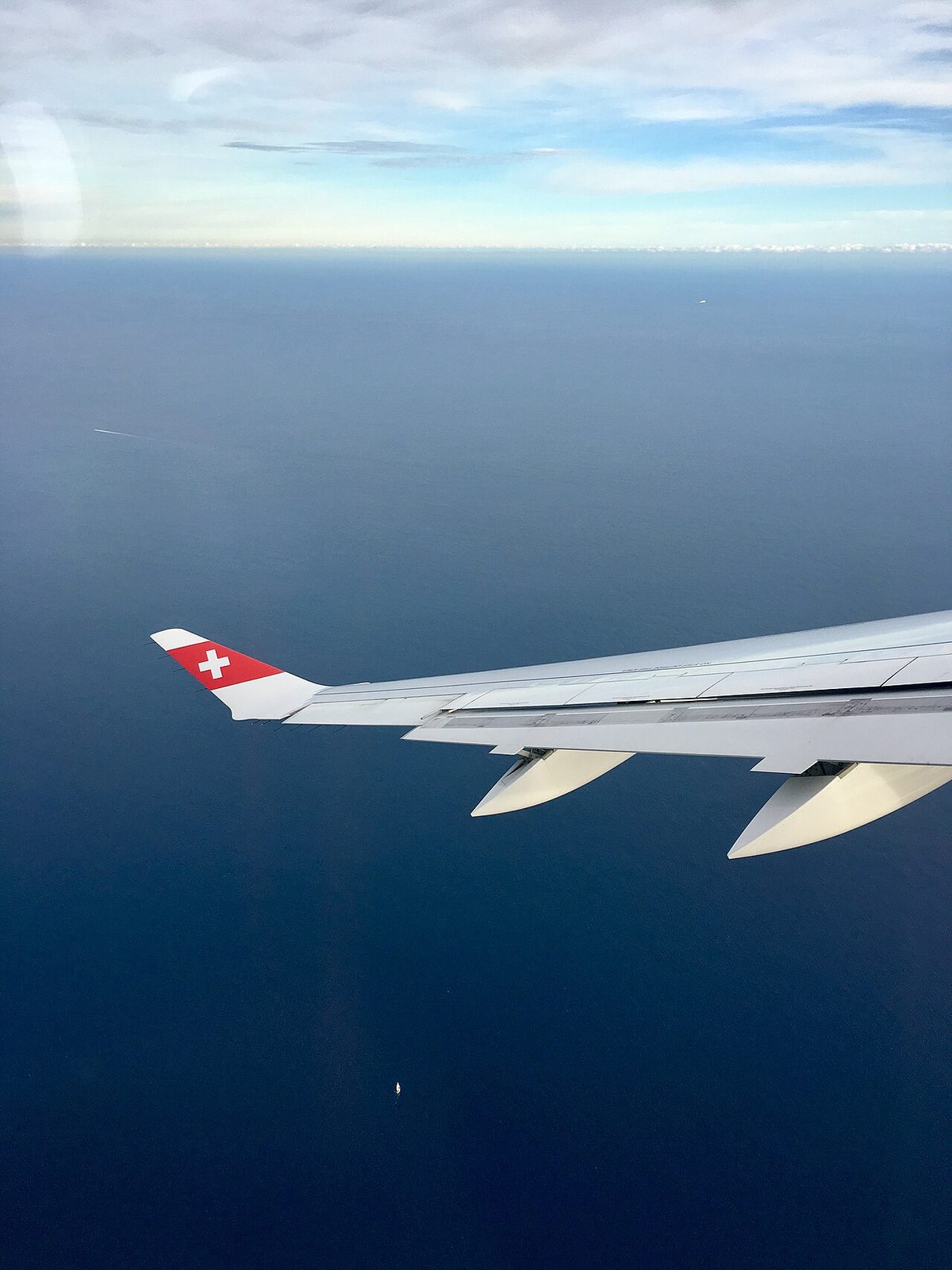 View from a cabin window of an airliner's swept wing over the sea, the wingtip in red and white with a Swiss cross, two streamlined fairings under the trailing edge.