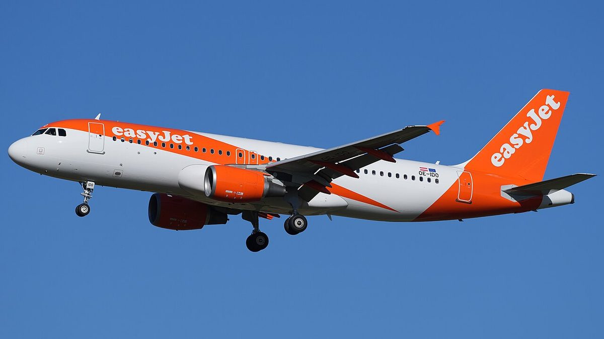 A white and orange easyJet Airbus A320 with landing gear down, flying against a clear blue sky.