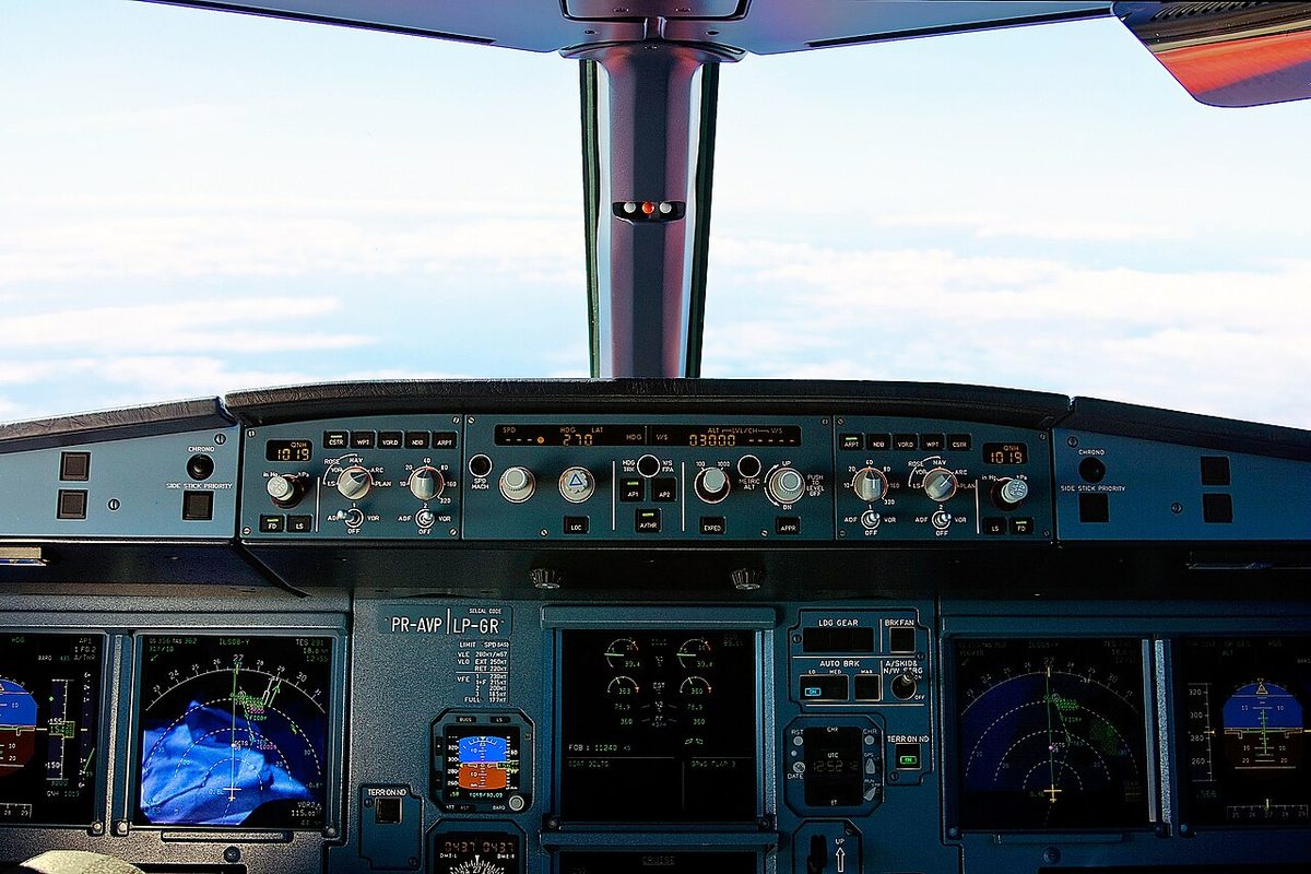 An Airbus A320 flight deck in flight: sky and clouds through the windscreen, the long Flight Control Unit panel of windows, knobs and pushbuttons across the glareshield, and lit display screens below it.