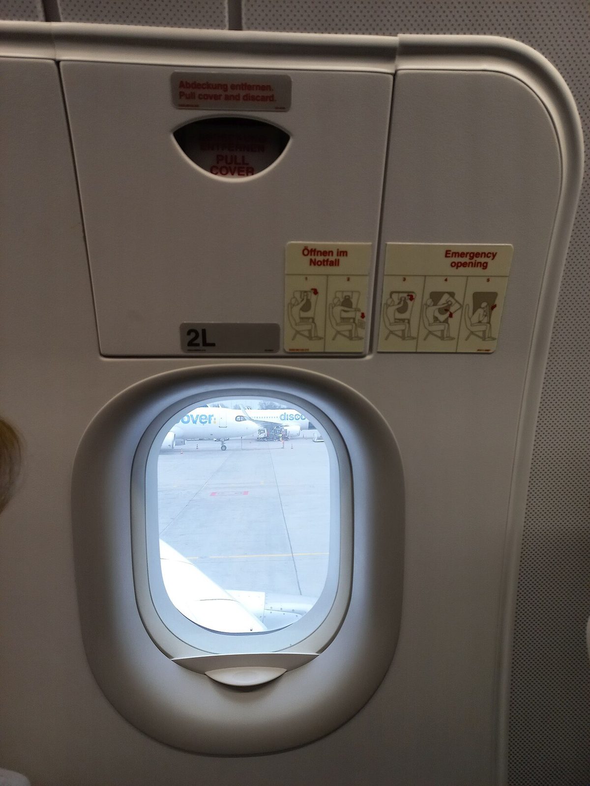 Inside an airliner cabin, an overwing exit hatch marked 2L, with a window, a cover marked PULL COVER at the top and two emergency opening placards in German and English.