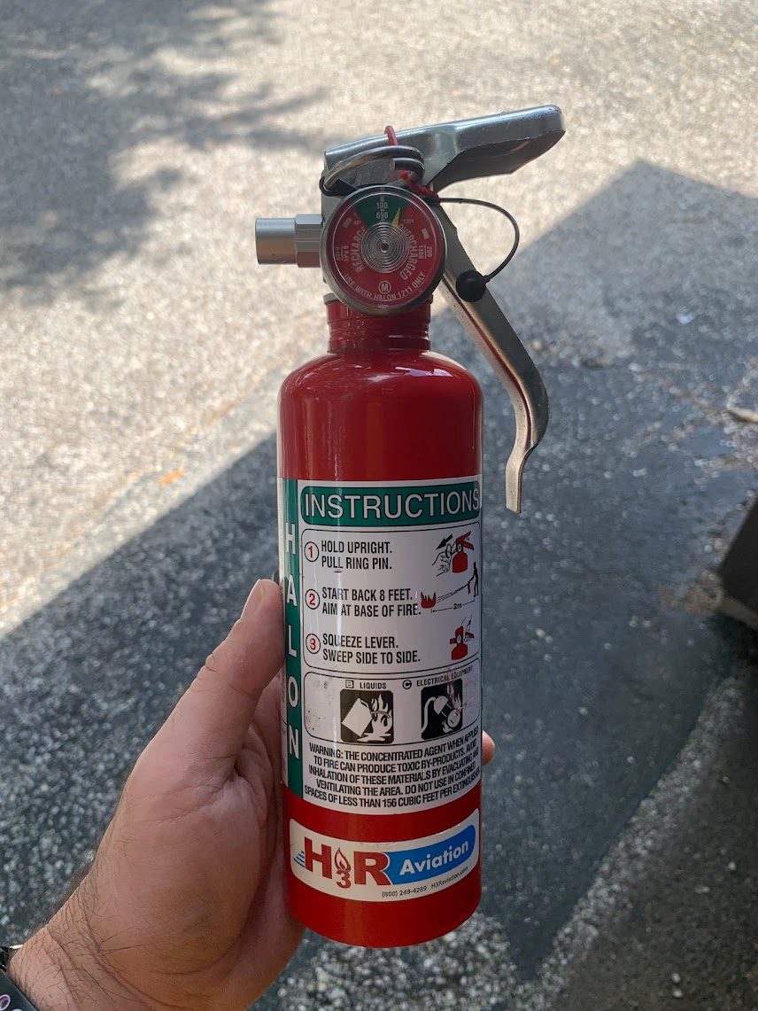 A hand holding a small red fire extinguisher with a pressure gauge, a black handle and an instruction label on the cylinder.