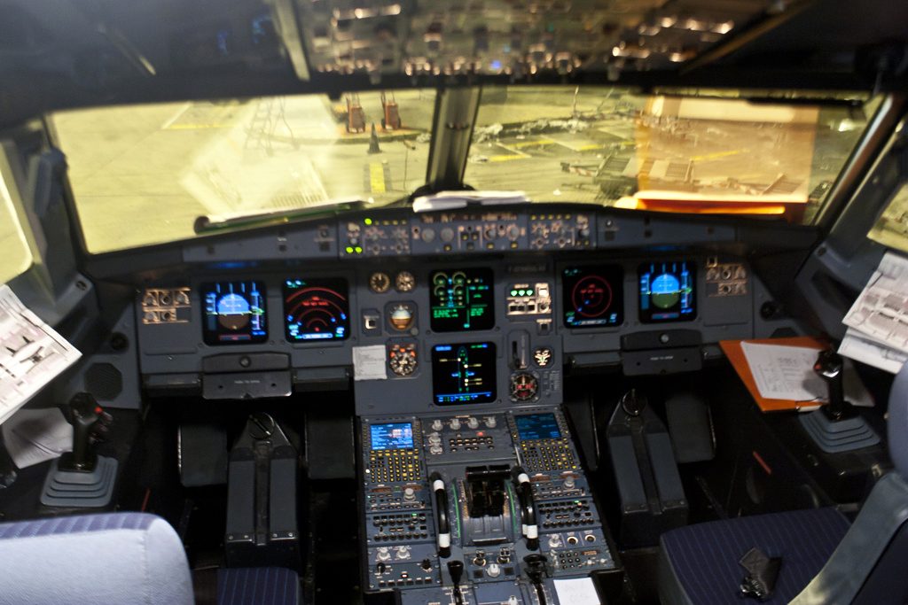 An Airbus A320 cockpit seen from behind the seats: six lit screens, the centre pedestal with thrust levers and a sidestick on each side console.
