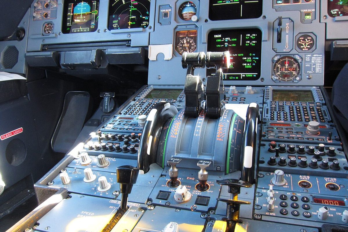 The centre pedestal of an airliner cockpit, with two thrust levers in the middle and a large dark wheel with a white mark on each side.