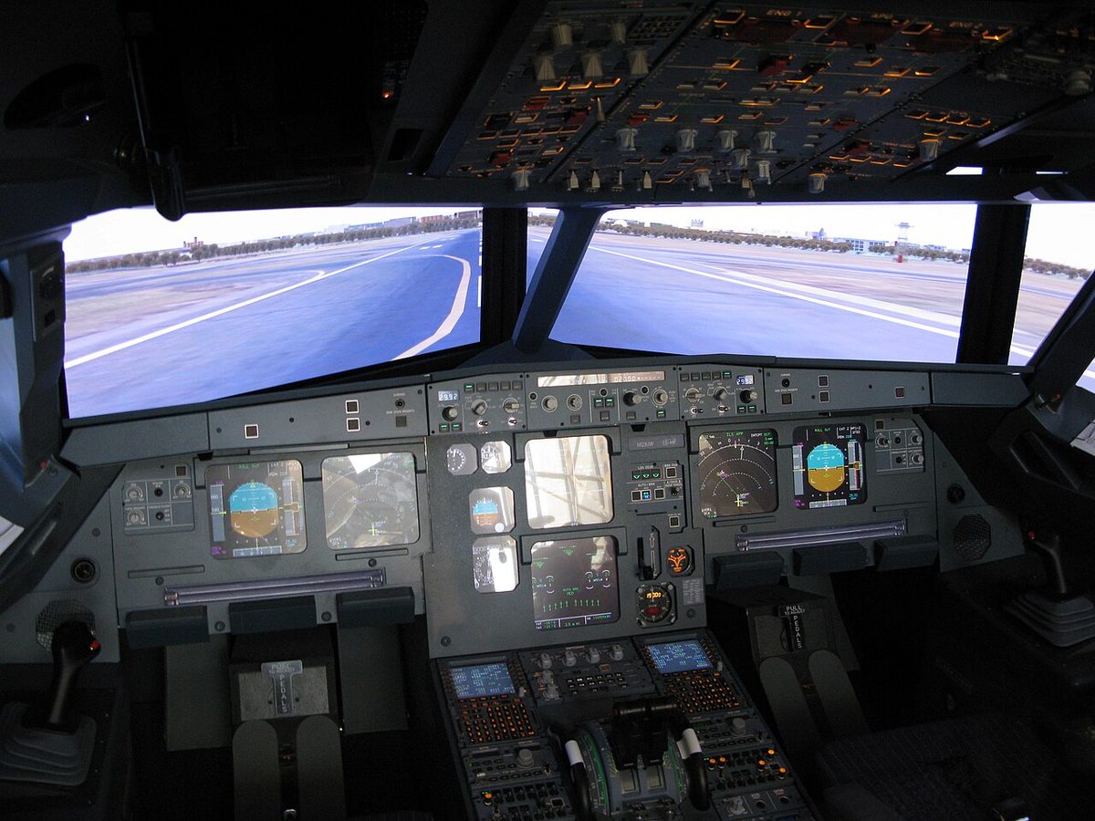 A dim airliner cockpit seen from behind the seats, with lit displays and a projected runway filling the windows.