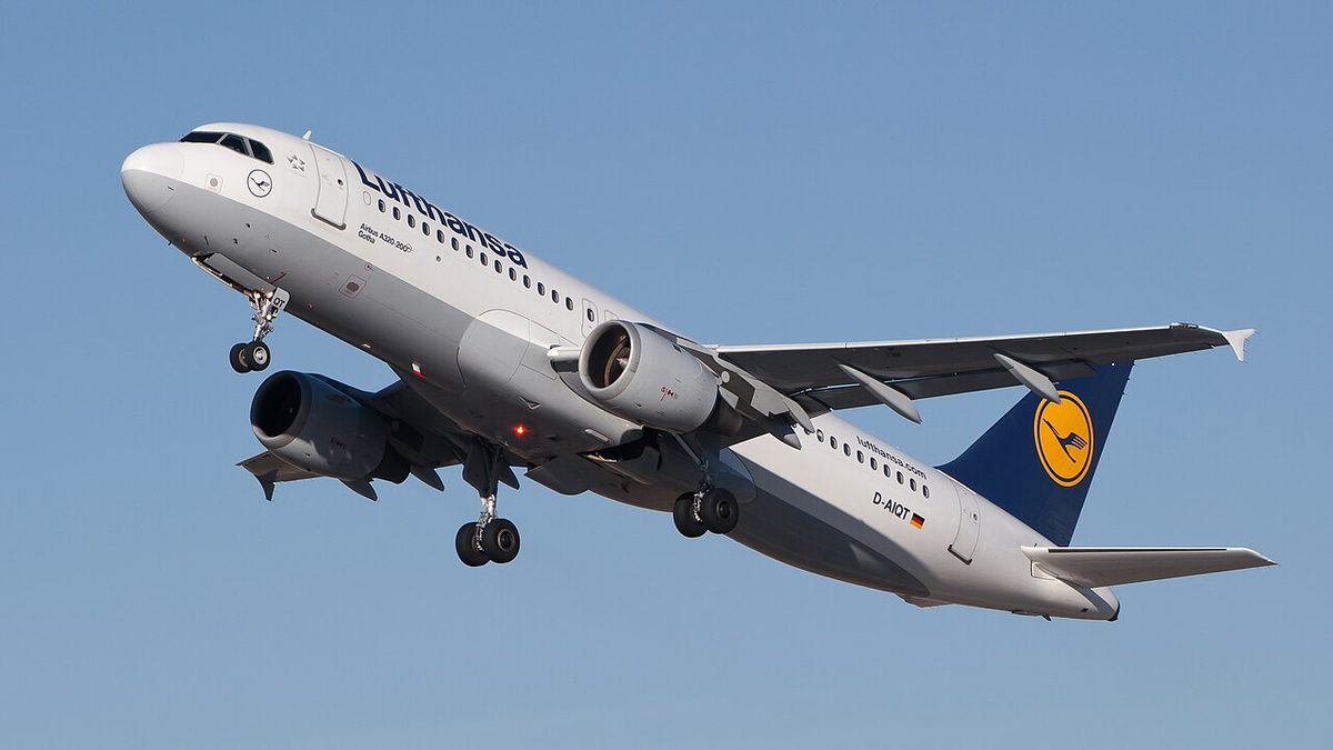 A white Lufthansa Airbus A320 just after lift-off, nose high and landing gear still down, against a blue sky.