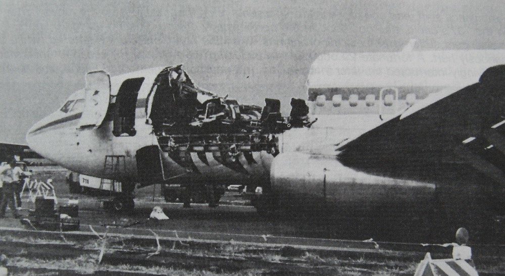 Black and white photo of an airliner on the ground, a long section of fuselage roof and upper walls missing behind the cockpit, exposing seats.
