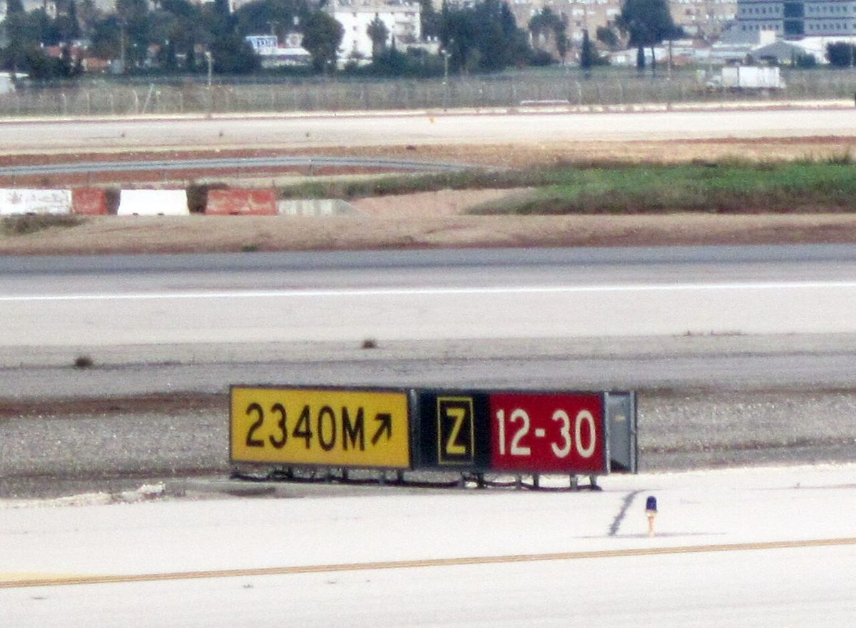 Airport signs beside a taxiway: a yellow sign reading 2340M with an arrow, a black sign with a yellow Z, and a red sign reading 12-30.
