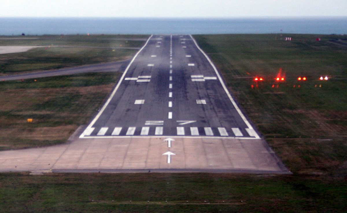 Final approach view of a runway with white threshold stripes and a row of four lights beside it on the right, three red and one white.