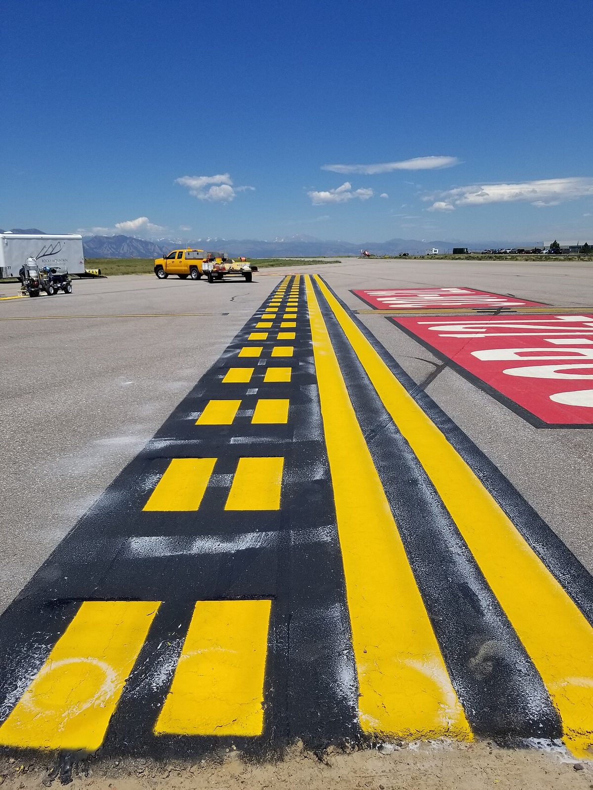 Close view along a freshly painted yellow marking, two solid and two dashed lines outlined in black, with a red painted sign on the right.