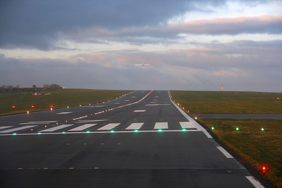 A runway at dawn seen from its end: white threshold stripes with a row of green lights along their near edge, runway edge lights on both sides and a small aircraft climbing away in the distance.