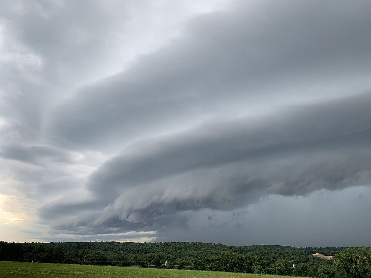A long, layered wedge of grey cloud sweeping low across the sky above a line of trees and a field.