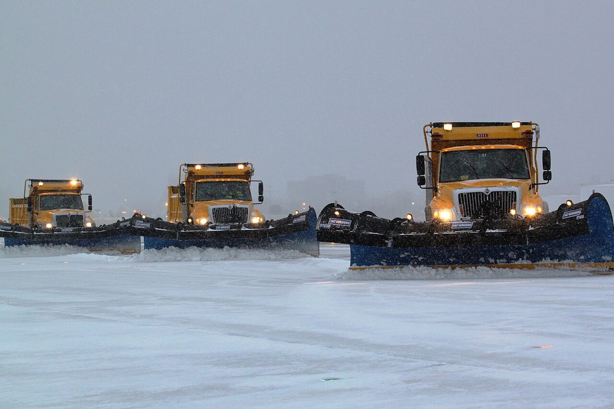 Three yellow snow ploughs with wide blades advancing side by side across a runway covered in snow.