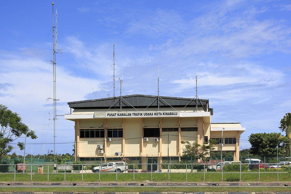 A low cream building signed Pusat Kawalan Trafik Udara Kota Kinabalu, with radio masts and aerials on its roof and a tall lattice mast beside it.