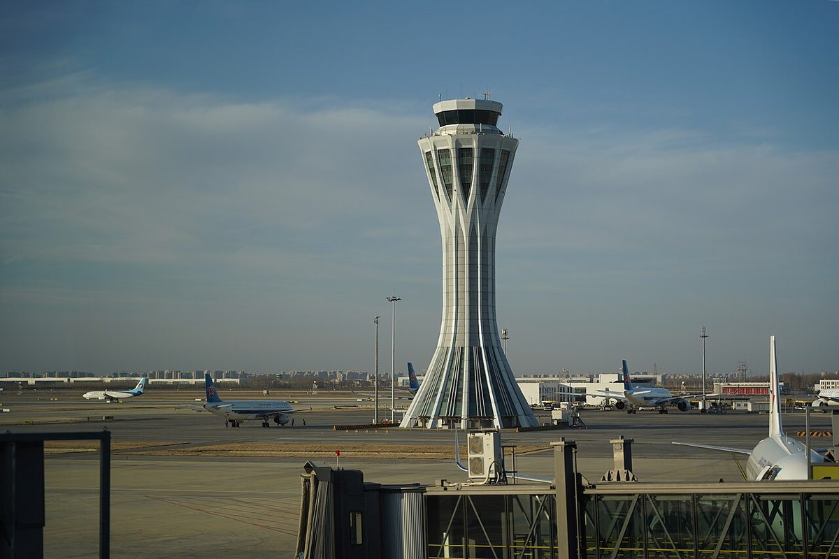 A tall white control tower with a glazed cab on top, standing on the apron of a large airport with airliners parked around it.