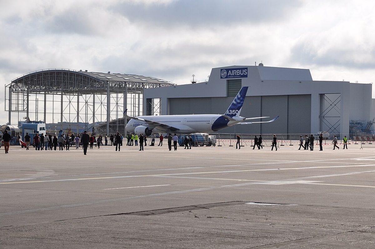 A large twin-engined Airbus airliner on the apron in front of Airbus hangars, watched by a crowd.