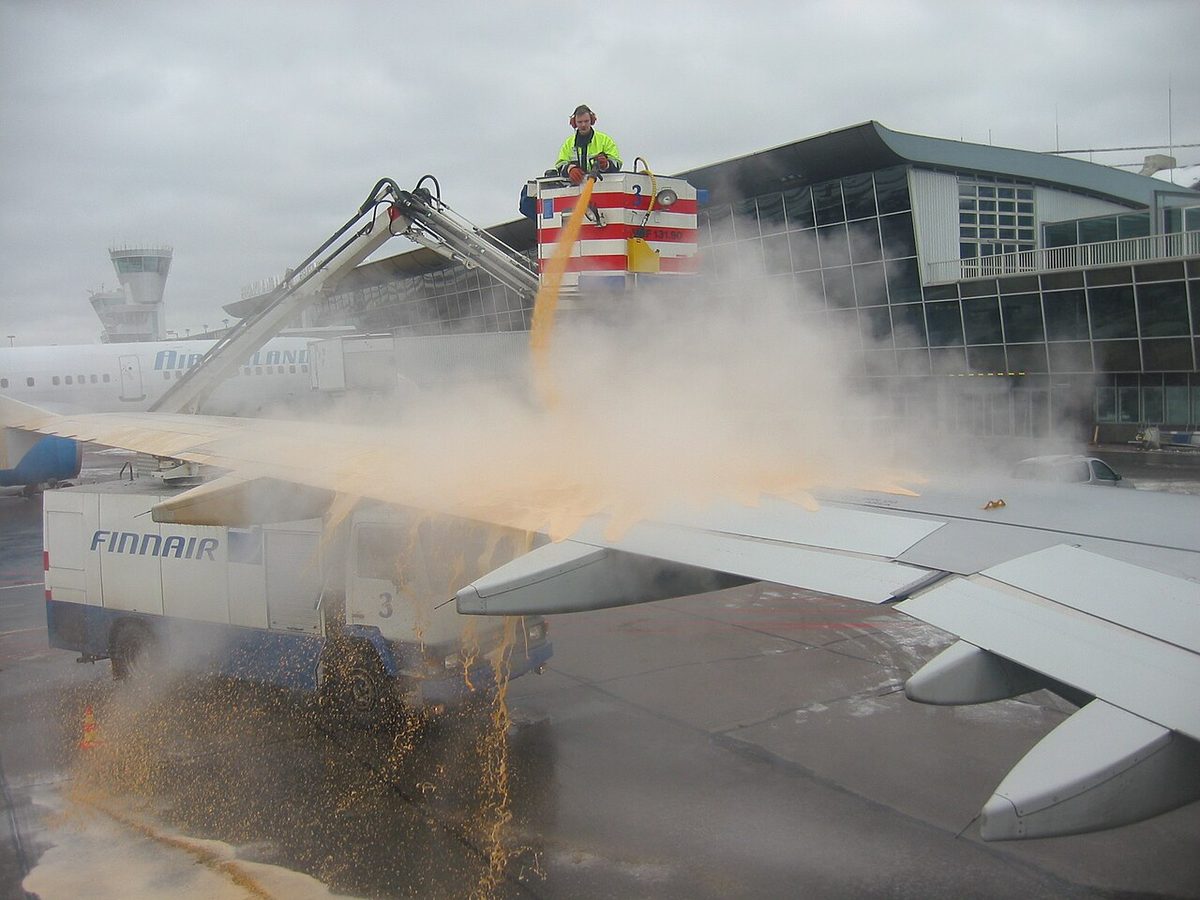 A worker in the raised basket of a truck sprays a heavy stream of orange-brown fluid onto an airliner's wing, a terminal building behind.