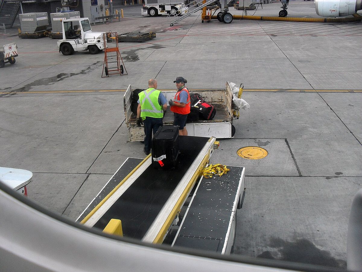 Seen from a cabin window, two ground handlers at a baggage cart beside a belt loader that carries a suitcase up to an airliner's hold.