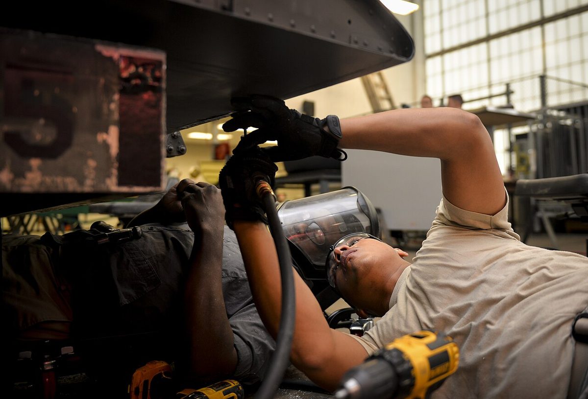 A technician in a face shield and gloves working with a drill on the underside of an aircraft structure in a hangar.