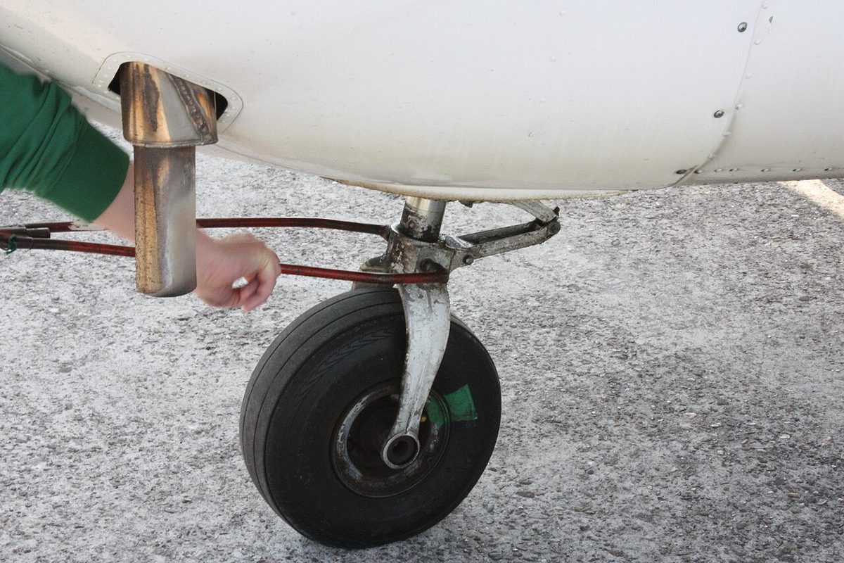 A small aircraft nose wheel on the ramp with a green painted mark running across the tyre sidewall and the wheel rim; an arm and a tow bar behind.