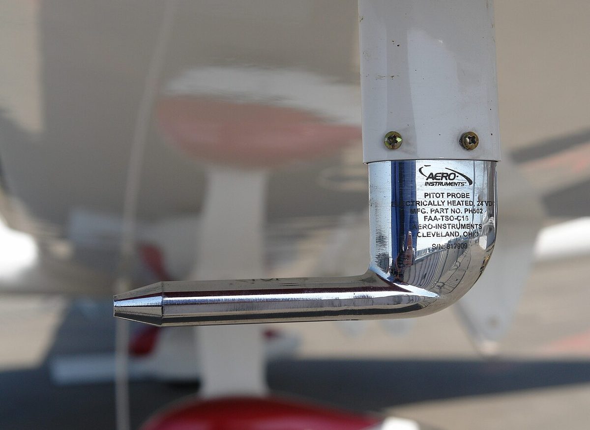 Chrome L-shaped pitot tube hanging below a white aircraft wing, its pointed open end facing forward into the airflow.