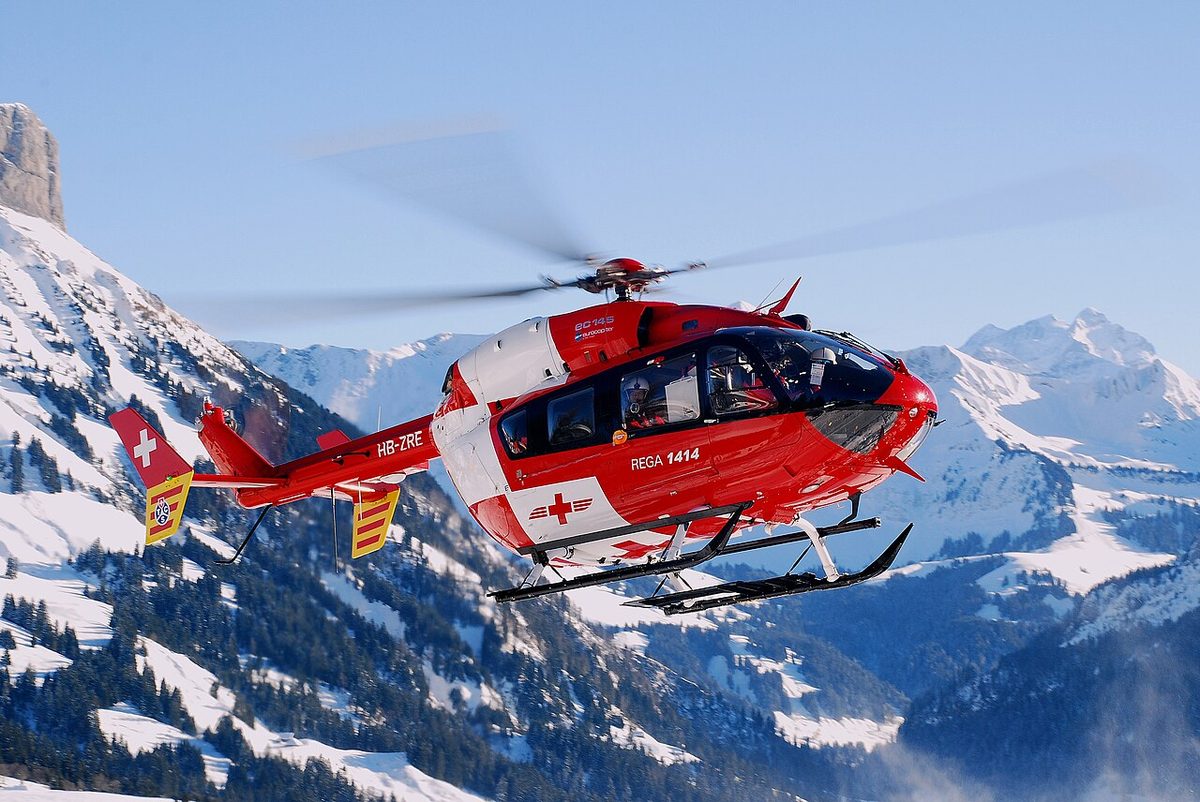 A red and white Rega air rescue helicopter in flight over snow-covered mountains.