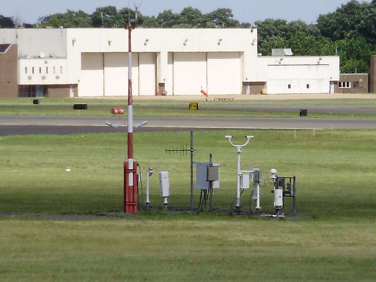 A cluster of weather sensors on poles and masts, including a red and white mast, on grass beside an airfield, with hangars behind.