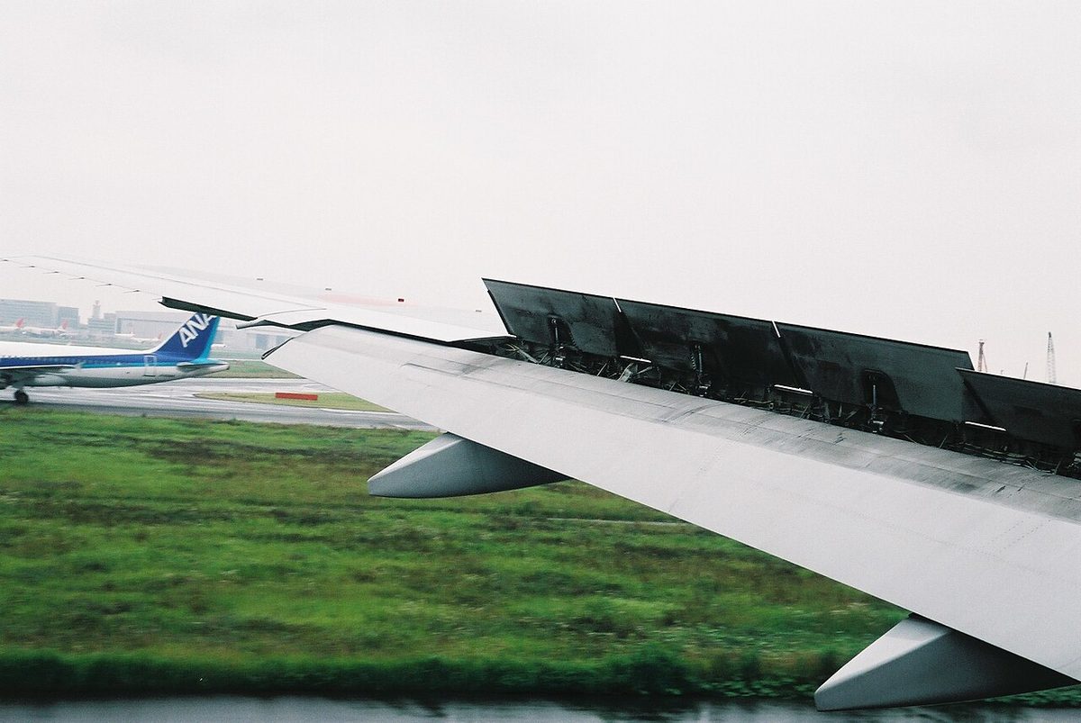 View from a cabin window of an airliner wing on the runway after landing, with a row of spoiler panels standing up from the upper surface.
