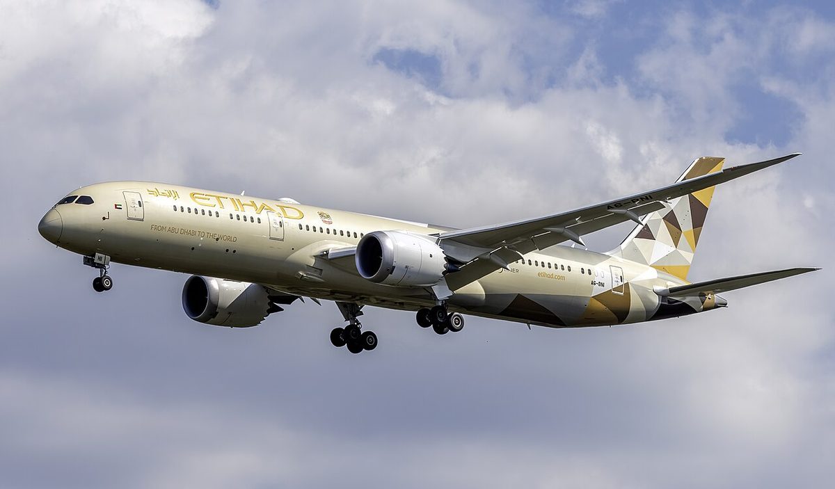 A beige and gold Etihad Boeing 787-9 on approach with its landing gear down, under white and grey cloud.
