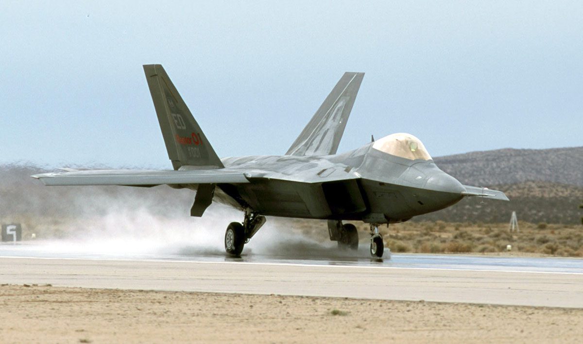 A grey fighter jet rolling along a wet runway, a cloud of spray rising from its main wheels.