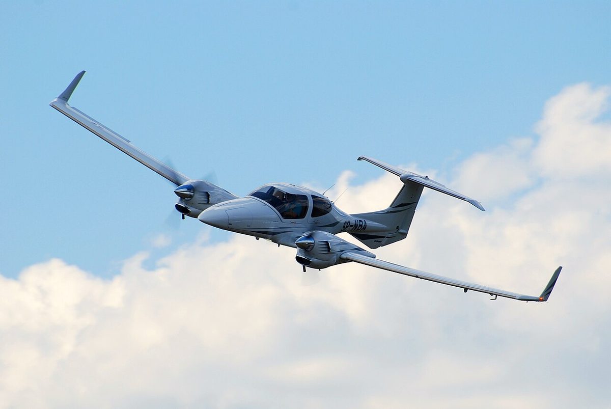 A white twin-engined light aeroplane with a T-tail and upturned wingtips flying past against a blue sky with white clouds.