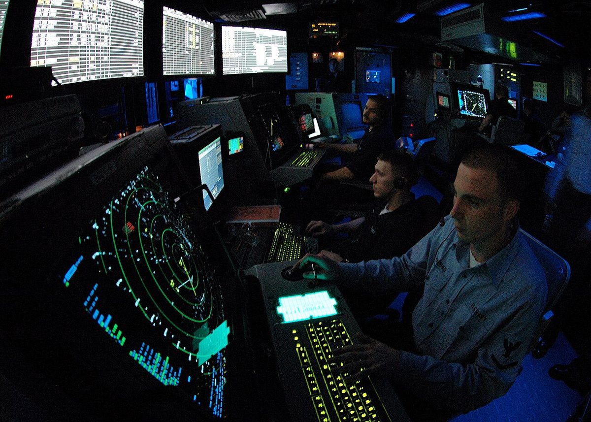 US Navy air traffic controllers at a row of radar consoles in a dimly lit room, a round radar display in the foreground and status screens above.