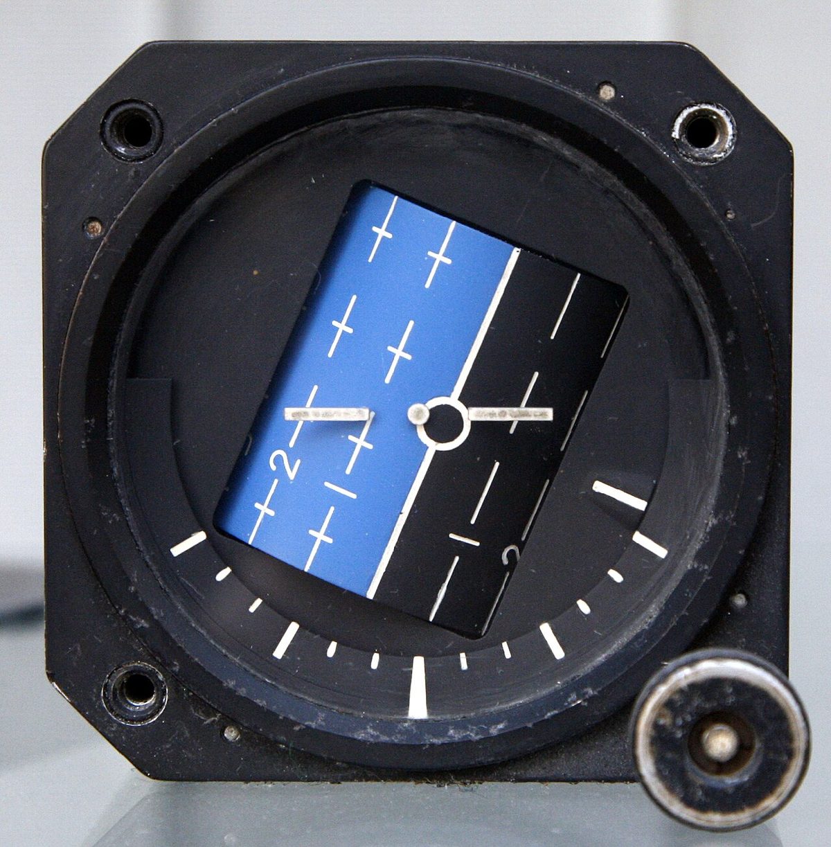 Attitude indicator with a blue and black horizon steeply tilted behind a fixed white aircraft symbol, bank marks around the bottom and a knob.