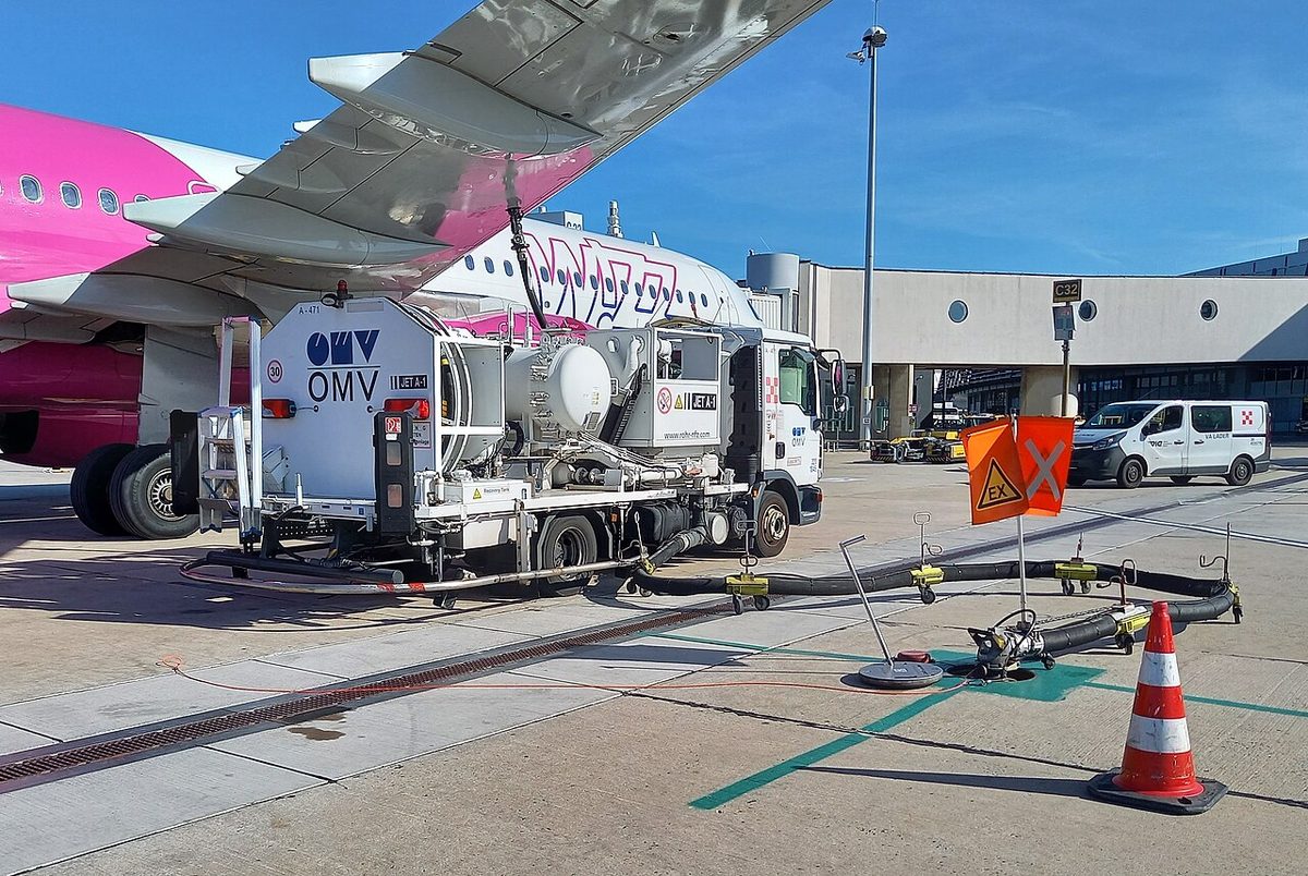 A fuel truck marked JET A-1 under an airliner's wing, a hose rising to the wing and another running across the apron to a ground point marked by a cone and flag.