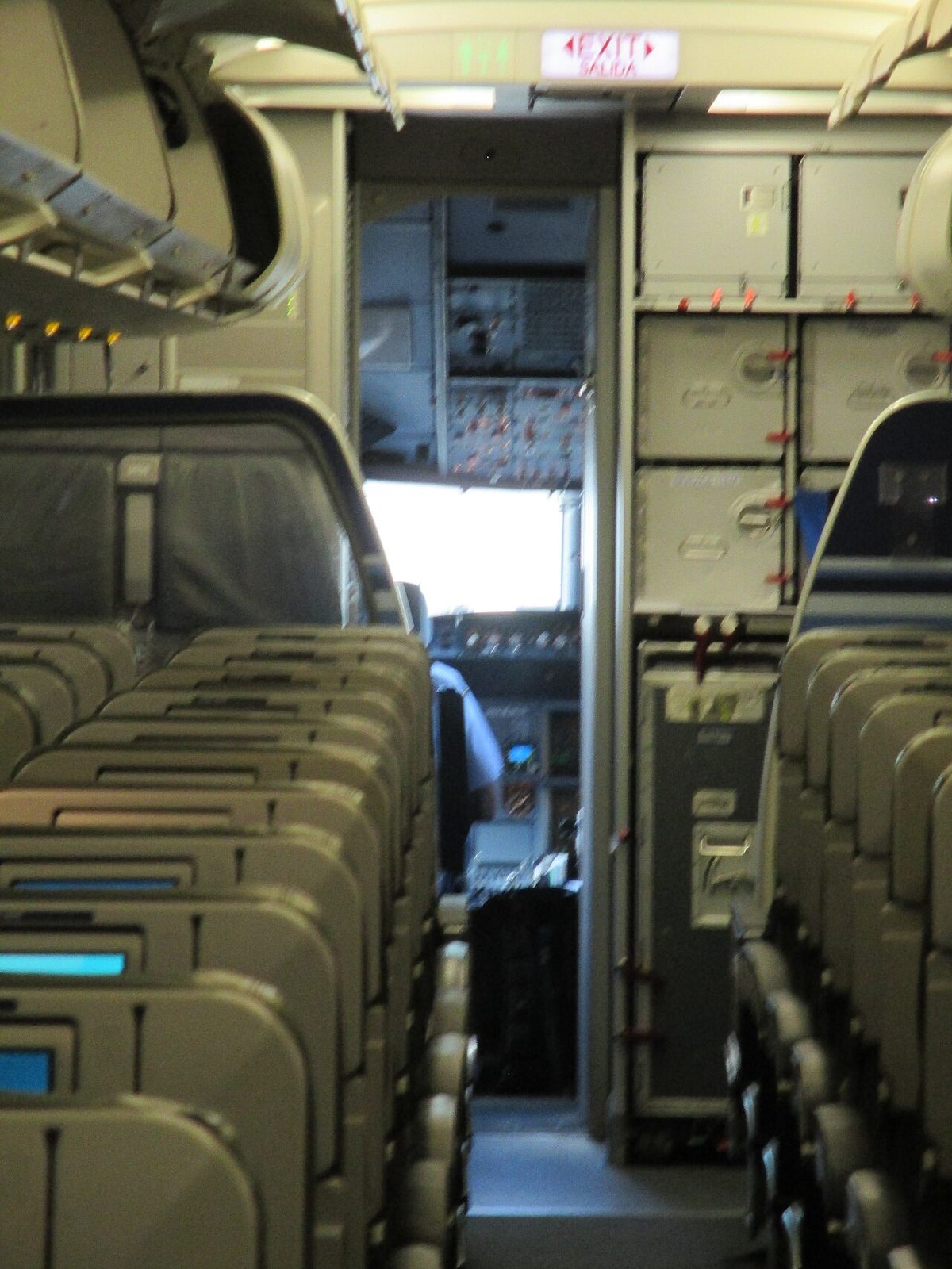 The flight deck of an Airbus A320 seen from the cabin aisle through the open flight deck door during boarding, with a pilot seated inside.