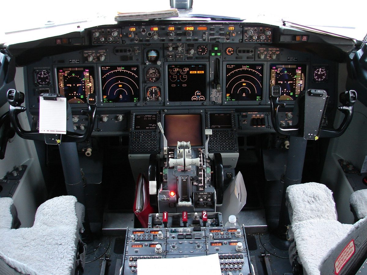Airliner flight deck seen from behind two sheepskin-covered seats, six display units lit across the main instrument panel, with the thrust levers on the control stand between the seats.