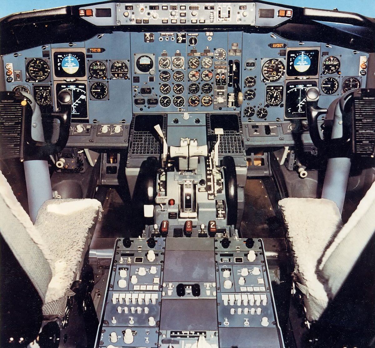 A Boeing 737-300 flight deck seen from behind the seats, with three fire switches marked 1, APU and 2 on the centre control stand just aft of the thrust levers.