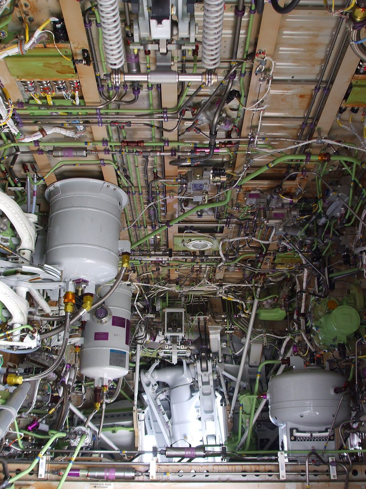 Looking up into an aircraft wheel bay crowded with pipes, cables and several white cylindrical tanks.
