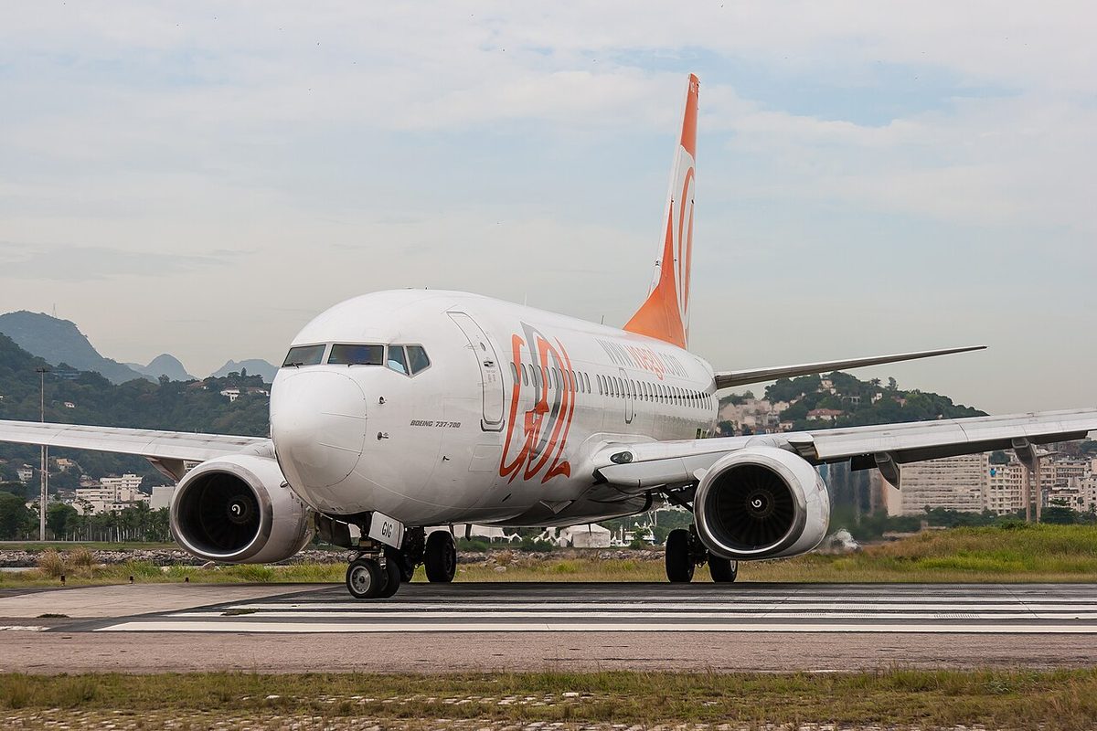 A white airliner with an orange tail and orange lettering on a runway, seen from the front, its two-wheel nose gear turned, with green hills and city buildings behind.