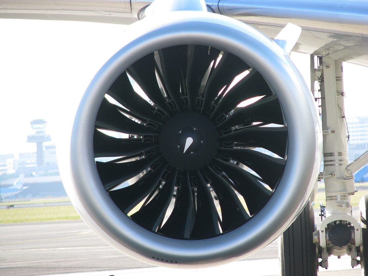 Front view of a large jet engine under a wing, a ring of curved dark fan blades inside a silver intake, with a main landing gear leg beside it.
