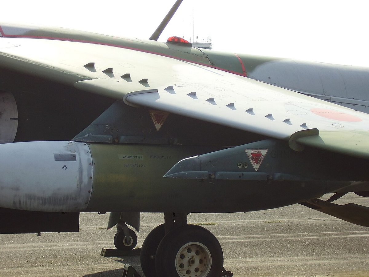 The upper surface of a camouflaged jet's swept wing, with rows of small triangular vanes standing up from the skin.