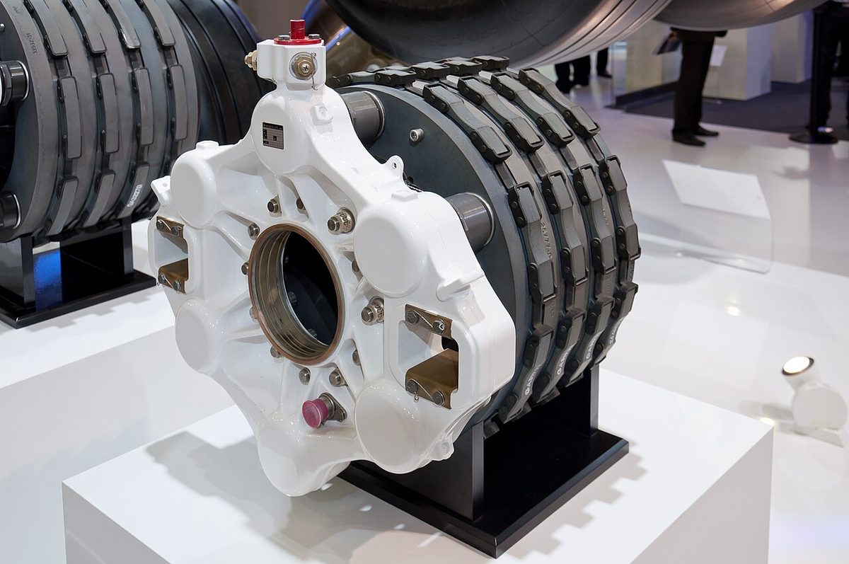 An aircraft wheel brake on a display stand: a white housing with pistons in front of a stack of dark grey carbon discs.