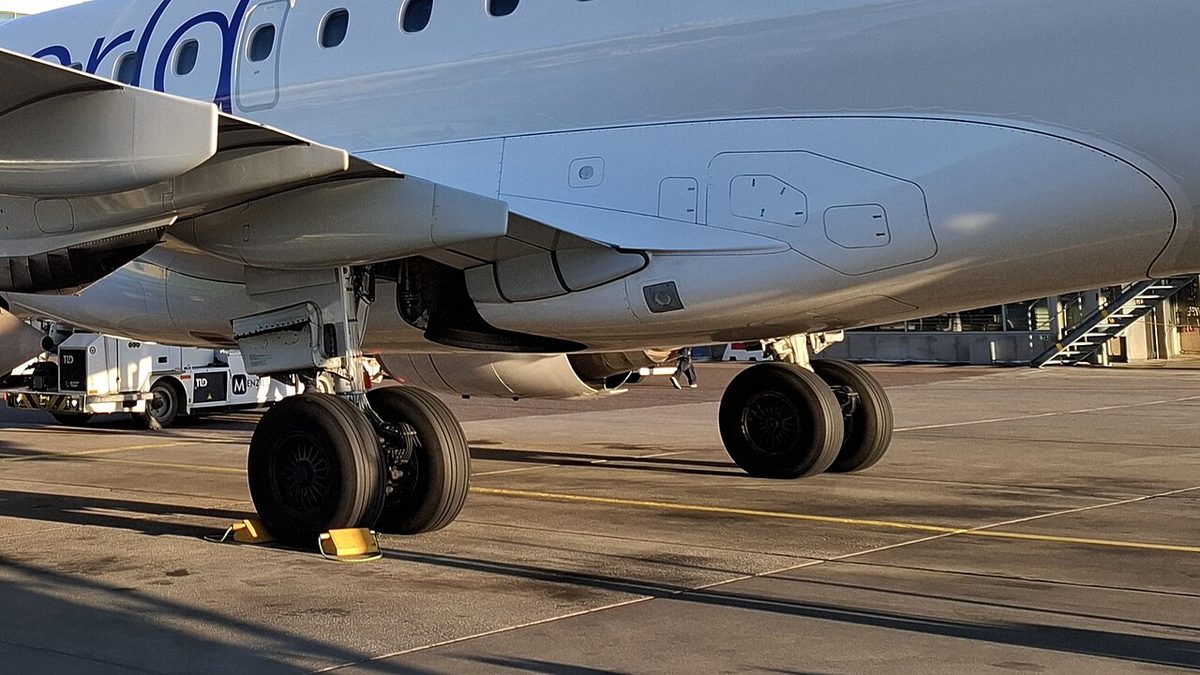 Underside of a parked regional jet showing both main landing gear legs, each with a pair of wheels, and a yellow chock on the apron.