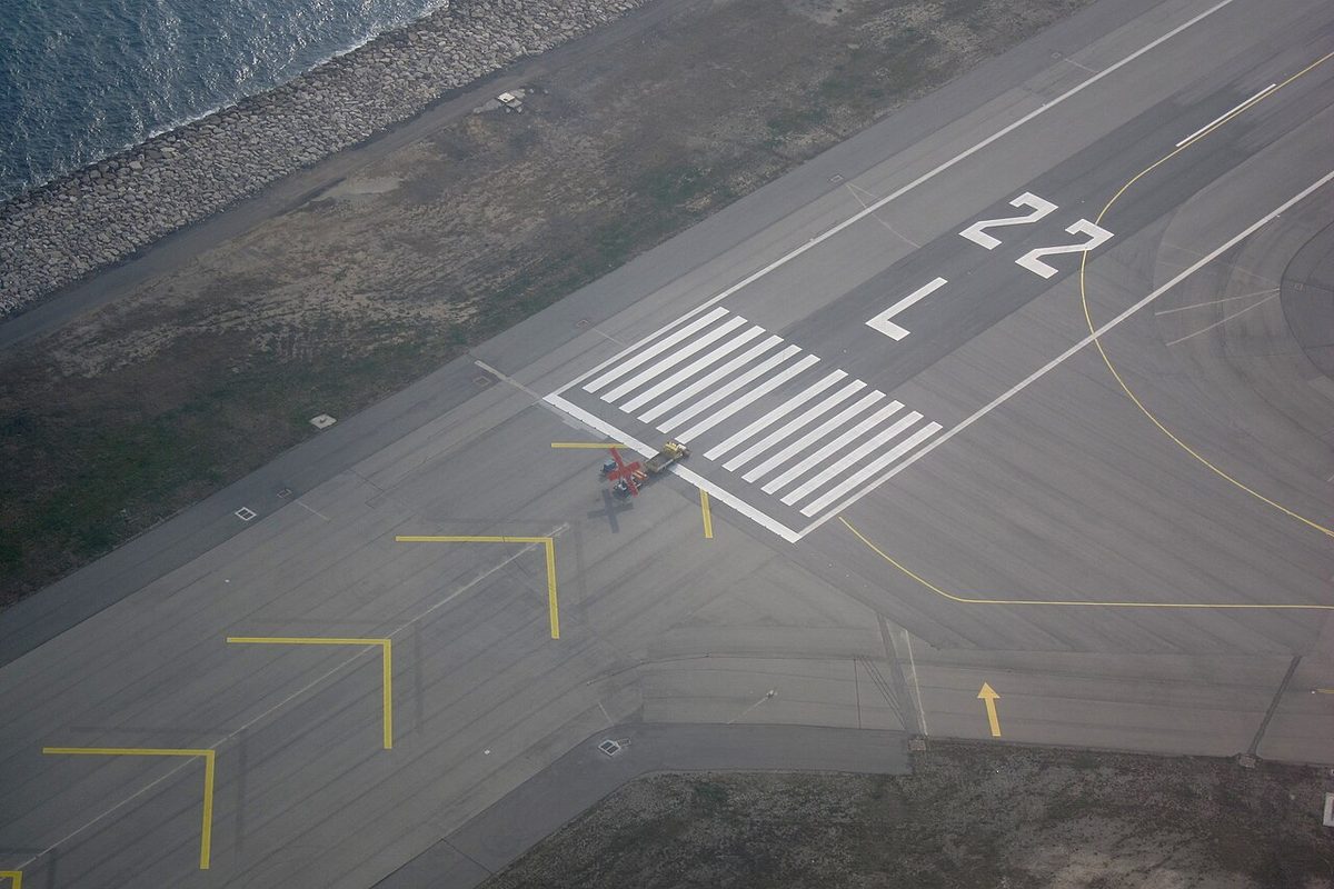 Aerial view of a runway end by the sea, with white threshold stripes, then a large L and the number 22, and yellow chevrons on the pavement before it.