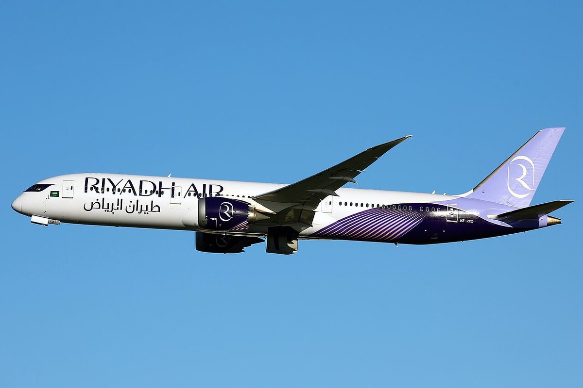 A white Riyadh Air Boeing 787 with a lilac tail and purple belly flying against a clear blue sky, landing gear retracted.