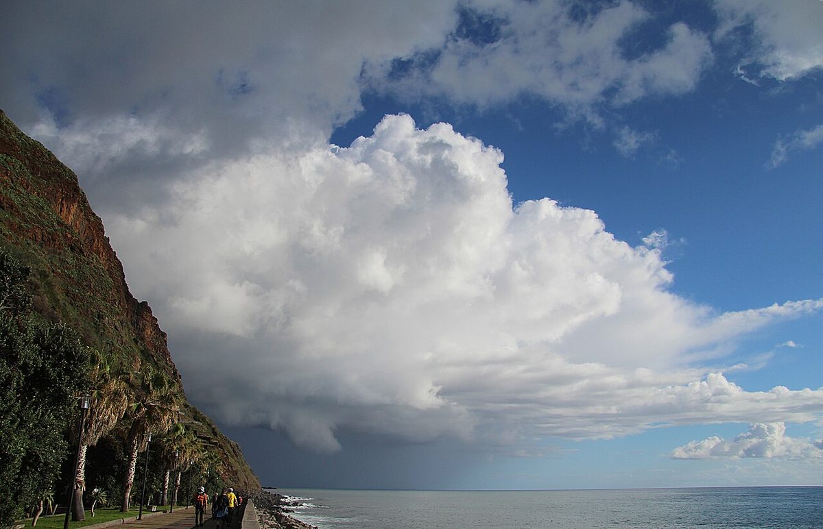 A towering white shower cloud over the sea beside a green cliff, with a grey curtain of rain beneath it and blue sky to the right.