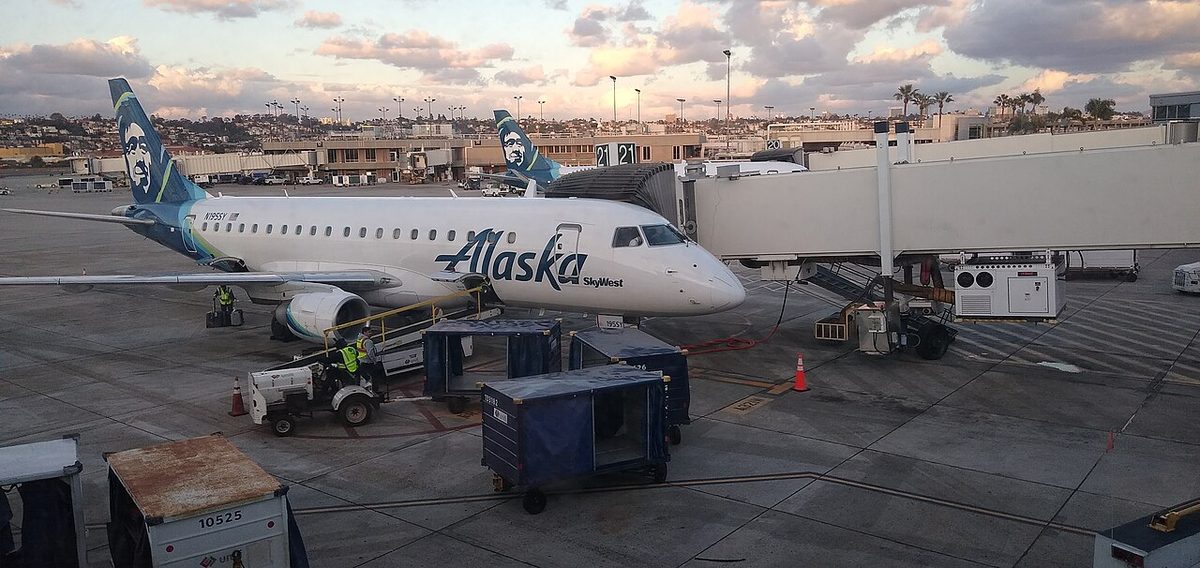 A white regional jet with a blue Alaska tail at a jet bridge at dusk, with baggage carts on the apron in front.